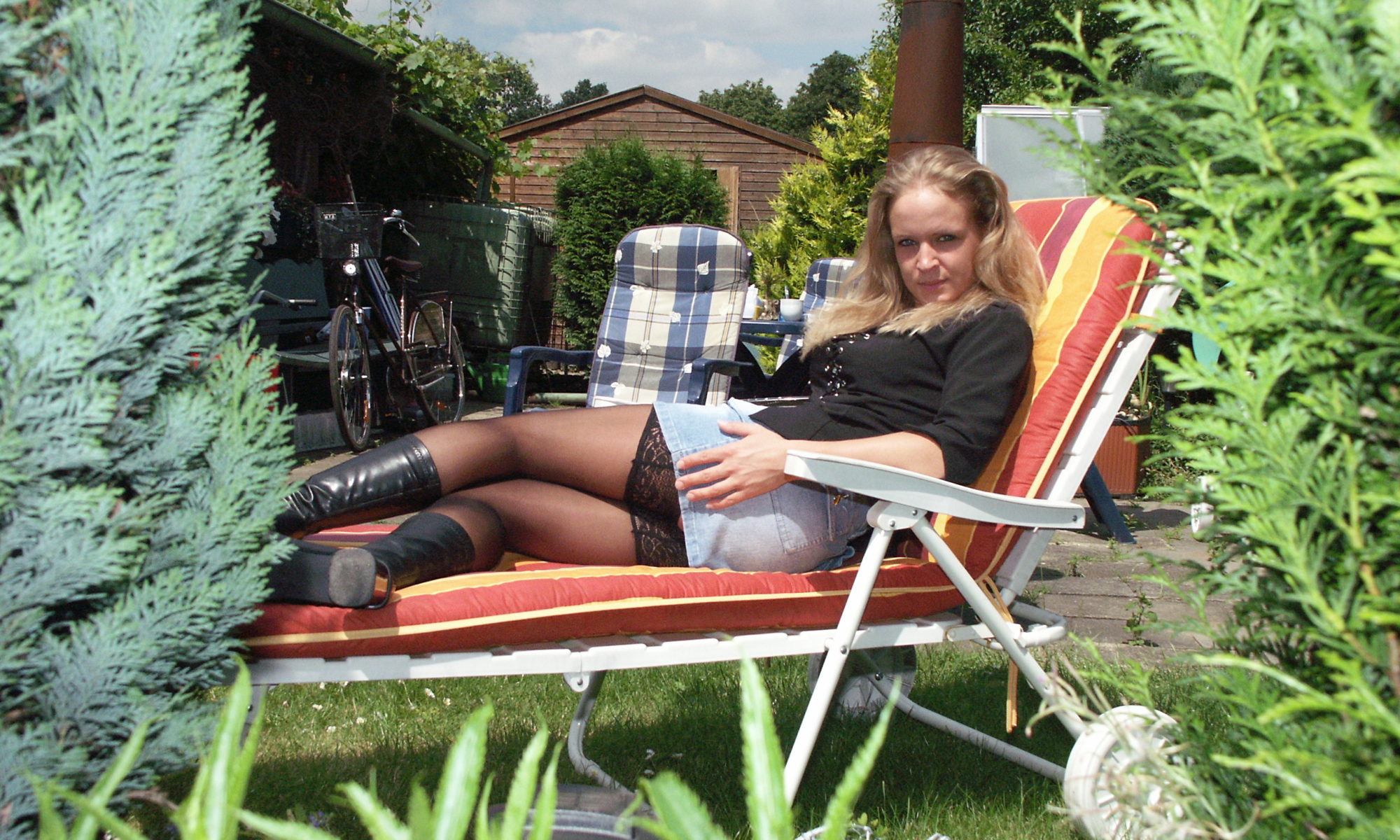 Junge Blondine in Minirock mit Strumpfhosen und hohen Stiefeln liegt erotisch auf einem Liegestuhl im Garten